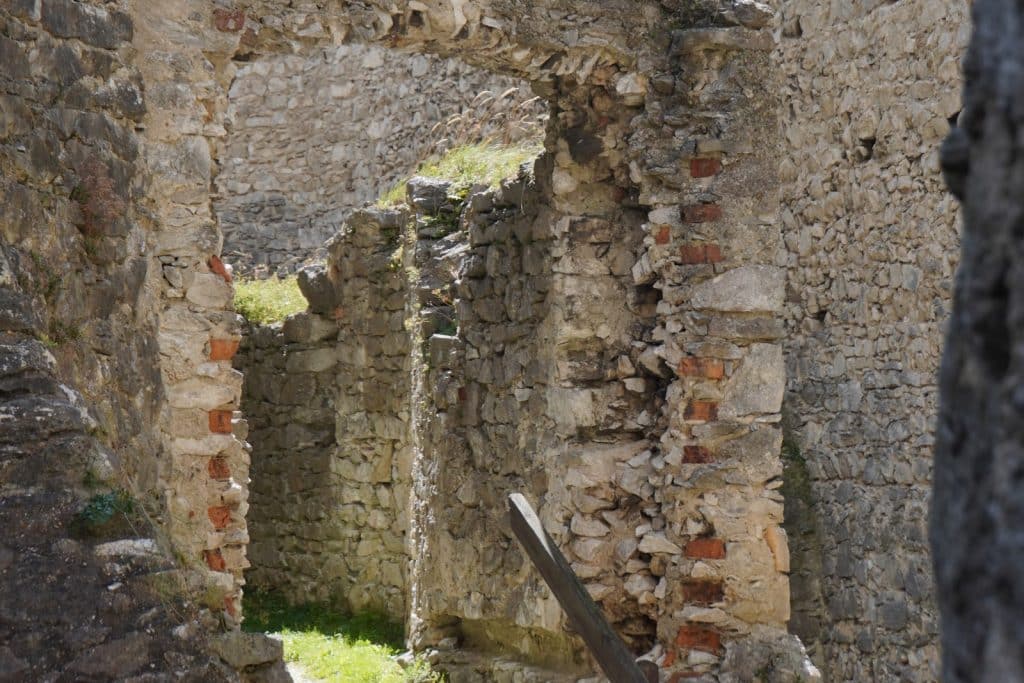 Blick durch einen steinernen Torbogen auf die verfallenen, hellen Bruchsteinmauern der Burg Rabí in Tschechien mit vereinzelten roten Ziegelsteinen und Grasbewuchs.