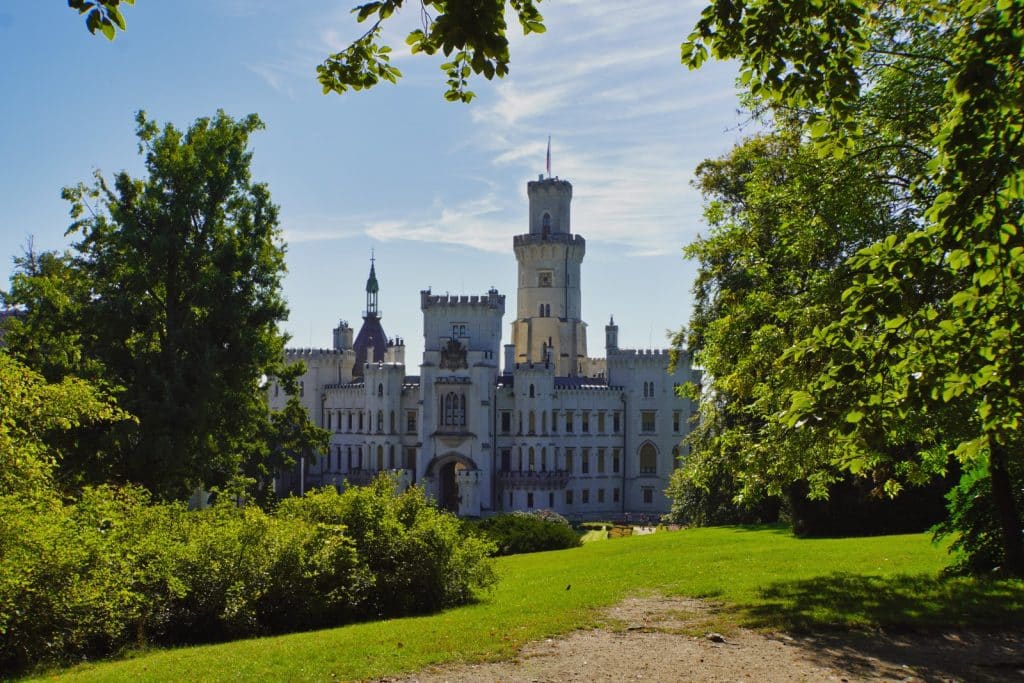 Das weiße, neugotische Schloss Hluboká in Tschechien mit seinen markanten Zinnen und Türmen thront hinter einer grünen Parkanlage unter strahlend blauem Himmel.