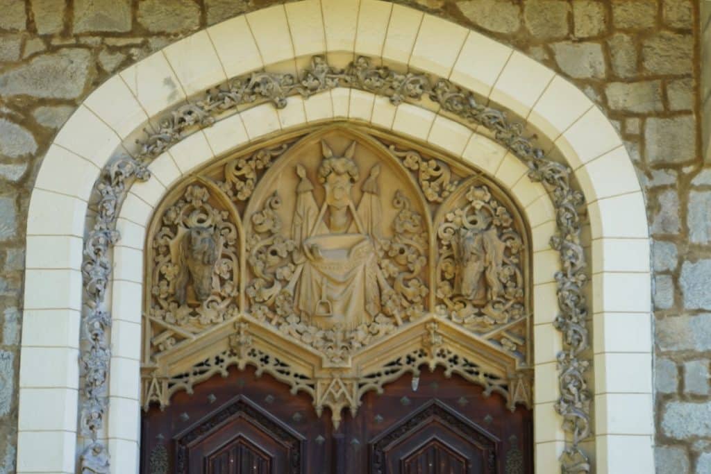 Detailreiche Sandsteinschnitzerei über einer Holztür von Schloss Hluboká in Tschechien, die ein zentrales Wappen mit Ritterhelm sowie zwei flankierende Pferdeköpfe in gotischen Bögen zeigt.
