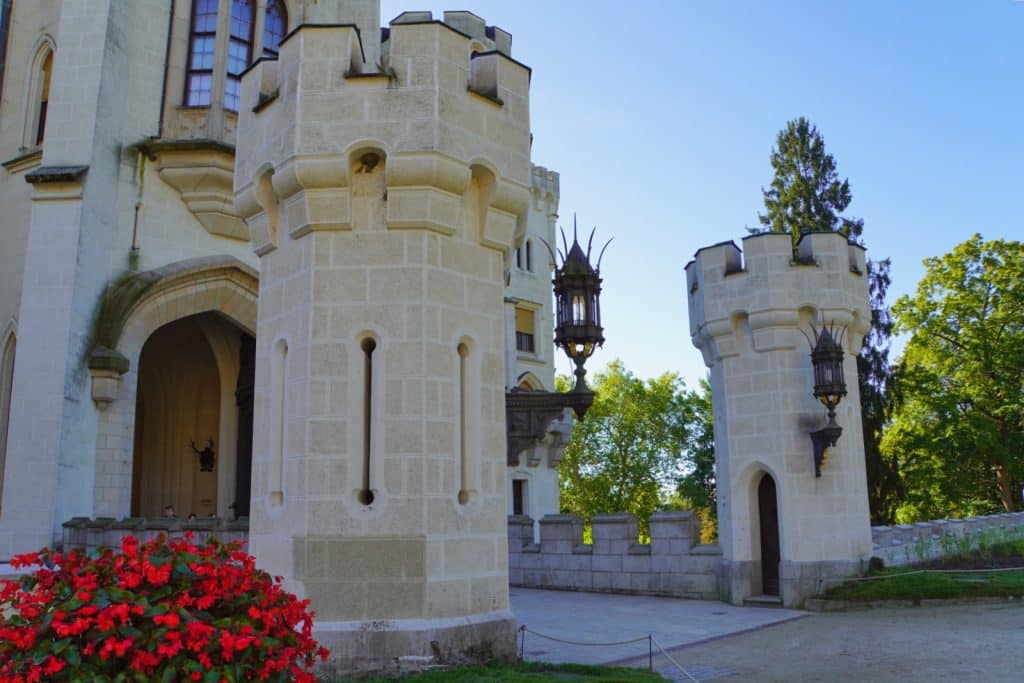 Die hellen, neugotischen Wehrtürme und Zinnen am Eingang von Schloss Hluboká in Tschechien ragen vor einem blauen Himmel auf, ergänzt durch rote Blumen im Vordergrund und schmiedeeiserne Laternen.