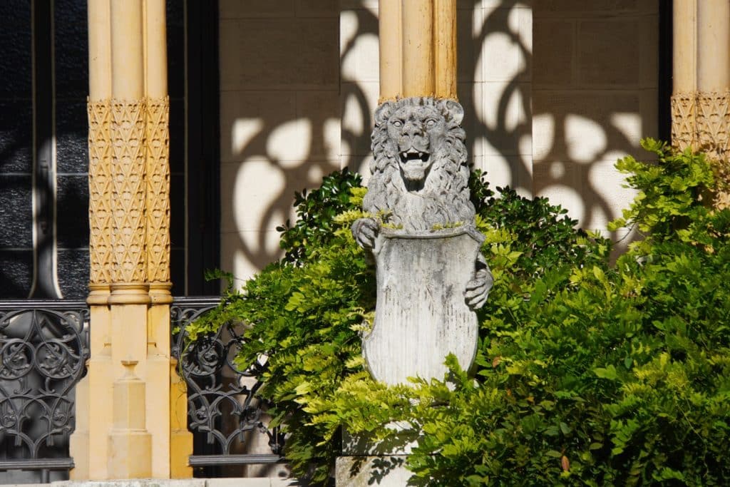 Eine steinerne Löwenstatue mit weit geöffnetem Maul hält ein Wappenschild auf einer Veranda von Schloss Hluboká in Tschechien, umrahmt von grünen Sträuchern und verzierten Säulen.