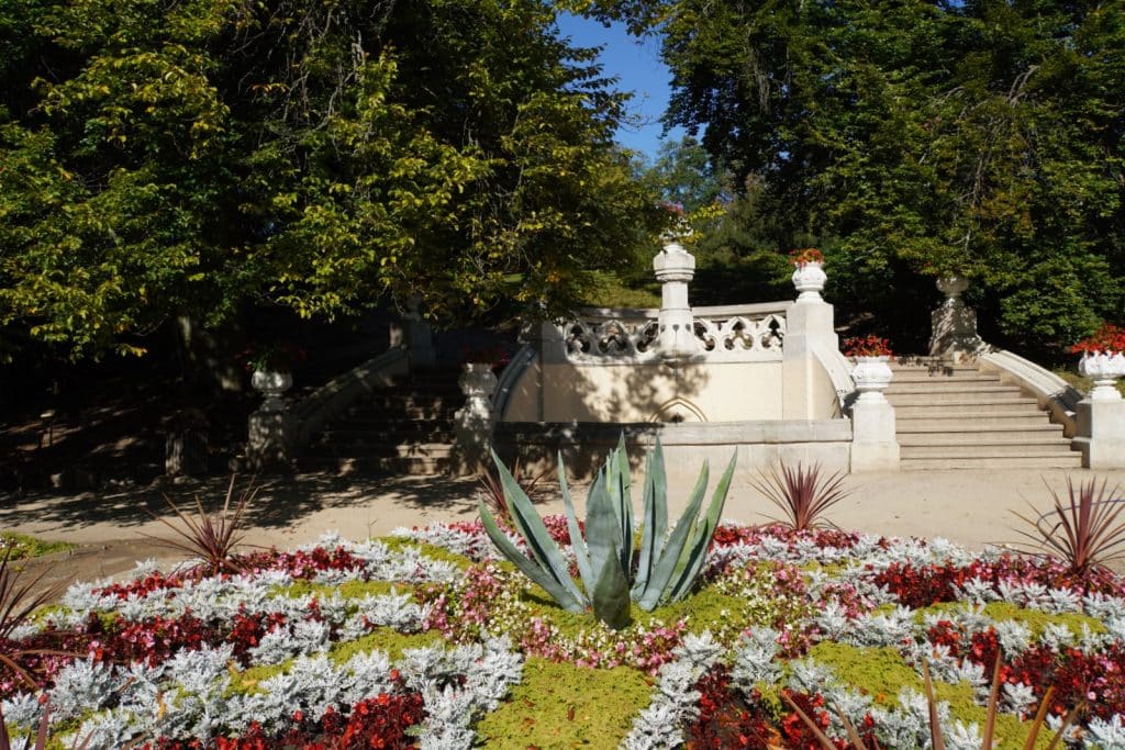Ein kreisförmiges Blumenbeet mit einer zentralen Agave und bunten Blüten im Vordergrund des Schlossparks von Hluboká in Tschechien, hinter dem eine weiße neugotische Treppenanlage zu bewaldeten Anhöhen führt.