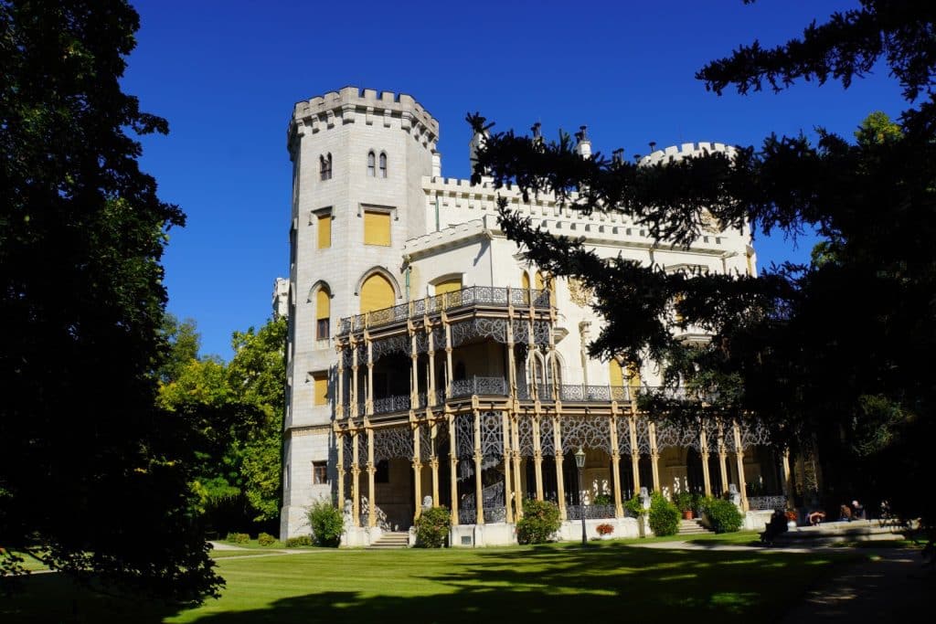 Die Rückseite des Schlosses Hluboká in Tschechien präsentiert sich als prachtvoller weißer Bau im neugotischen Stil mit einem markanten Zinnenturm und einer filigranen, mehrstöckigen Eisenkonstruktion der Veranda vor einer gepflegten Rasenfläche.