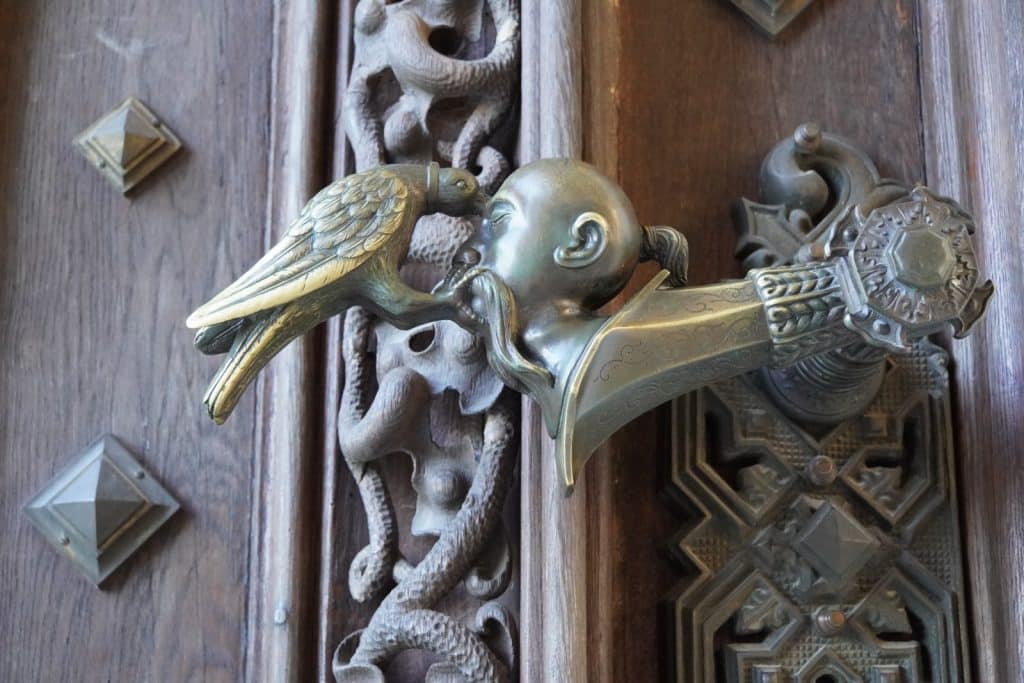 Eine detailreiche bronzene Türklinke am Schloss Hluboká in Tschechien zeigt einen Raben, der einem abgeschlagenen Kopf mit langem Schnurrbart und Zopf ins Auge pickt. Das kunstvolle Metallelement ist an einer dunkelbraunen, geschnitzten Holztür mit geometrischen Beschlägen angebracht.