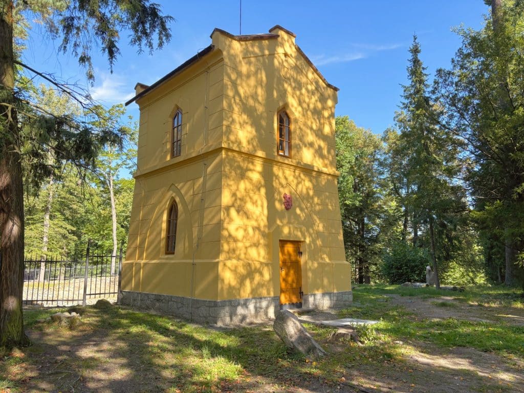 Ein zweistöckiger, gelber Wasserturm im neugotischen Stil mit spitzbogigen Fenstern und einer hölzernen Eingangstür steht auf dem Gelände von Schloss Hluboká in Tschechien, umgeben von Bäumen und einem schmiedeeisernen Zaun.