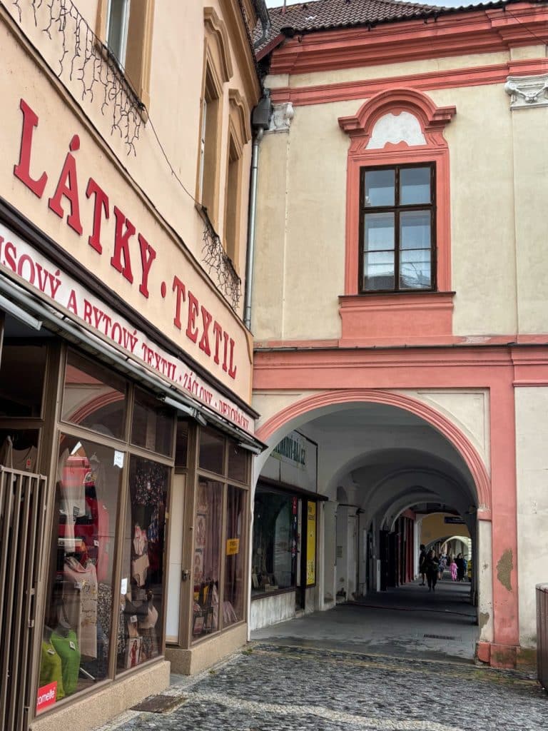 Ein historischer Arkadengang in Žatec mit einem Textilgeschäft im Vordergrund und einem Blick durch die gewölbten Durchgänge der gepflasterten Gasse.