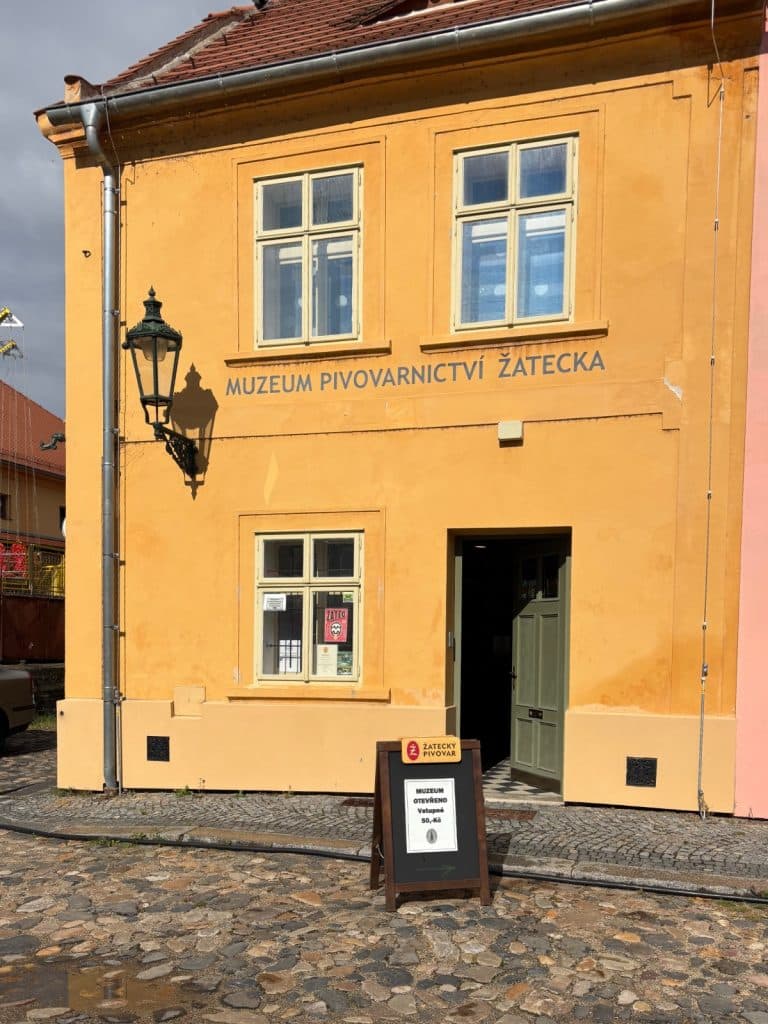 Die Außenansicht des zweistöckigen, ockerfarbenen Gebäudes des Biermuseums in Žatec zeigt den Eingangsbereich mit einem Hinweisschild auf dem Kopfsteinpflaster. Über dem Erdgeschoss prangt der Schriftzug „MUZEUM PIVOVARNICTVÍ ŽATECKA“ unter zwei Sprossenfenstern.