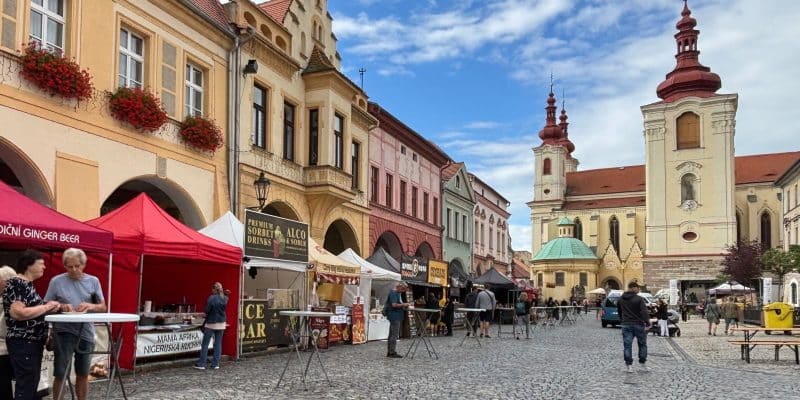 Das Hopfenfest in Žatec findet auf einem gepflasterten Marktplatz statt, der von historischen Gebäuden, Verkaufsständen mit roten Zelten und einer barocken Kirche im Hintergrund gesäumt wird. Besucher schlendern zwischen den Ständen und Stehtischen unter einem leicht bewölkten blauen Himmel.