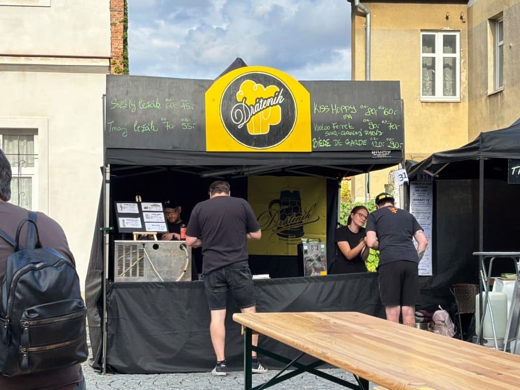 Ein belebter Bierstand der Brauerei Dráteník auf dem Hopfenfest in Žatec, an dem Personal hinter dem Tresen Kunden bedient und eine handgeschriebene Kreidetafel verschiedene Biersorten und Preise anzeigt. Im Vordergrund stehen ein hölzerner Biertisch und ein Besucher mit Rucksack, während im Hintergrund historische Gebäude zu sehen sind.