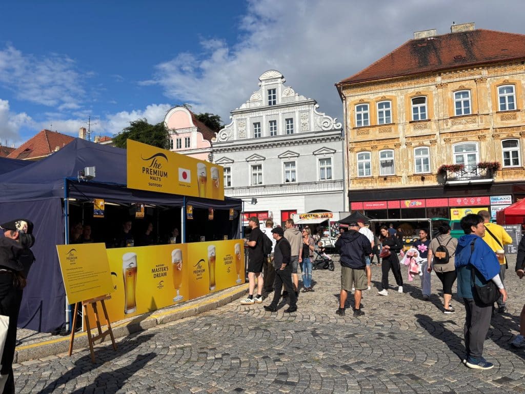 Ein gelber Bierstand von Suntory The Premium Malt's auf dem Hopfenfest in Žatec, umgeben von Menschen auf einem gepflasterten Marktplatz vor historischen Gebäuden.