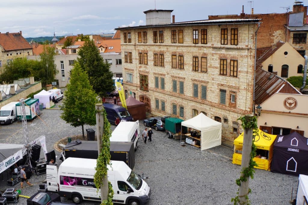 Vorbereitungen für das Hopfenfest in Žatec auf einem gepflasterten Platz mit weißen Transportern, Zelten und technischem Equipment vor einem historischen, mehrstöckigen Gebäude.