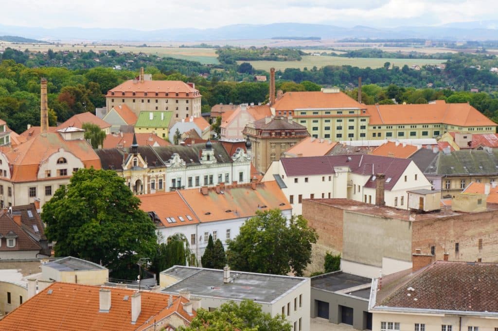 Blick über die Dächer der tschechischen Stadt Žatec mit ihren charakteristischen Hopfenmagazinen, die durch rote Ziegeldächer und hohe Schornsteine vor einer hügeligen Landschaft geprägt sind.