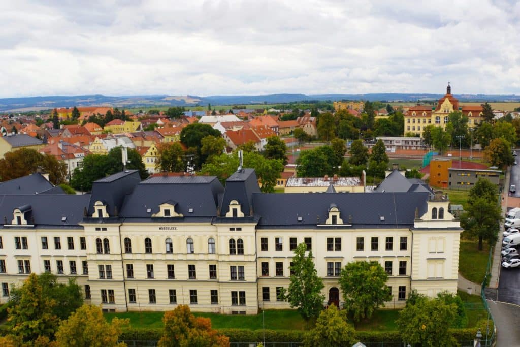 Blick vom Hopfenturm in Žatec auf ein großes, gelbes Schulgebäude mit dunklem Mansarddach vor dem Panorama der Stadt und der umliegenden Hügellandschaft.