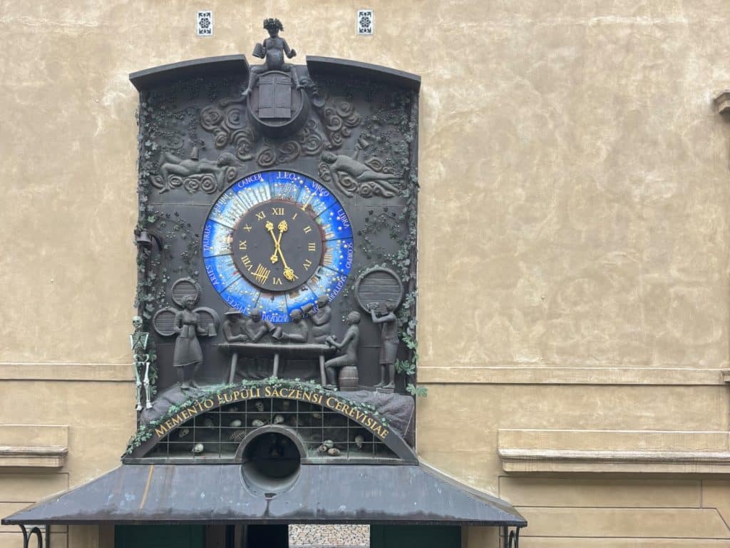 Die kunstvoll gestaltete Hopfenuhr in Žatec zeigt ein astronomisches Zifferblatt mit Tierkreiszeichen, umgeben von Bronzereliefs, die Szenen der Hopfenverarbeitung und des Biergenusses darstellen. Über dem Zifferblatt thront eine Figur, während darunter die lateinische Inschrift „Memento lüpüli saczensi cerevisia“ über einem Gitter mit Totenköpfen prangt.