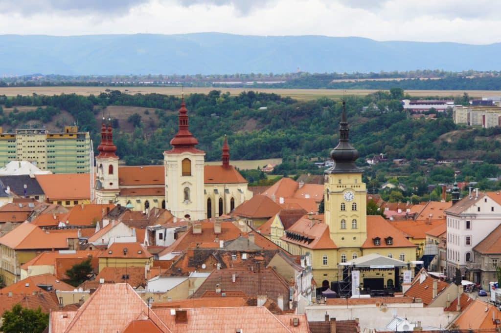 Blick über die roten Ziegeldächer der tschechischen Stadt Žatec auf das gelbe Rathaus mit seinem markanten Uhrturm und die dahinterliegende Kirche vor einer hügeligen Landschaft.