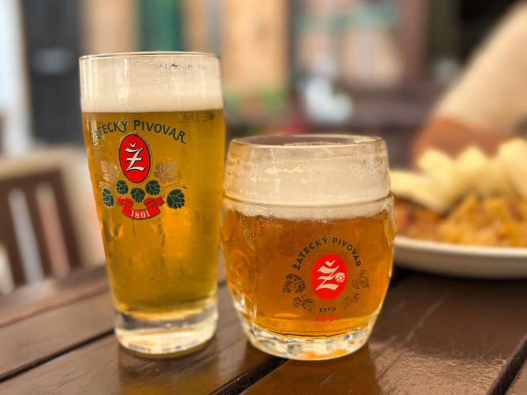 Zwei Gläser frisch gezapftes Bier der Marke Žatecký Pivovar stehen auf einem dunklen Holztisch in einem Restaurant in Žatec, im Hintergrund ist ein Teller mit Essen angedeutet.