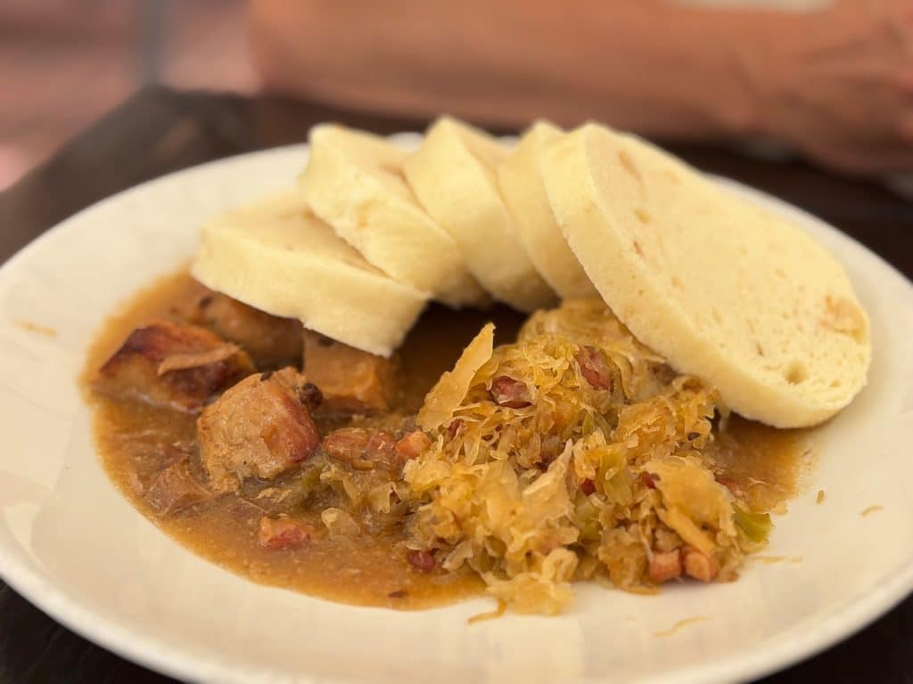 Ein weißer Teller mit einem traditionellen tschechischen Gericht aus einem Restaurant in Žatec, bestehend aus Schweinebraten in Soße, Sauerkraut und mehreren Scheiben böhmischer Knödel.