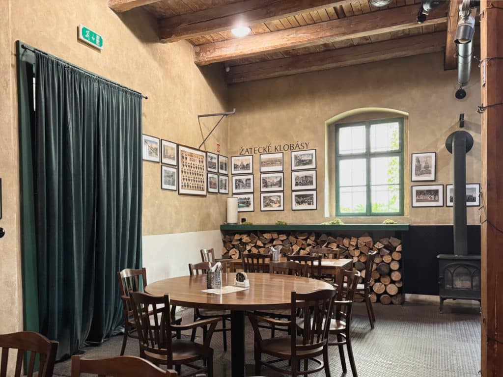 Der rustikale Innenraum eines Restaurants in Žatec zeigt einen runden Holztisch mit Stühlen vor einer Wand mit historischen Fotos, einem Holzstapel unter dem Fenster und einem gusseisernen Ofen. Ein schwerer grüner Vorhang hängt links neben dem Eingangsbereich unter einer Decke mit massiven Holzbalken.