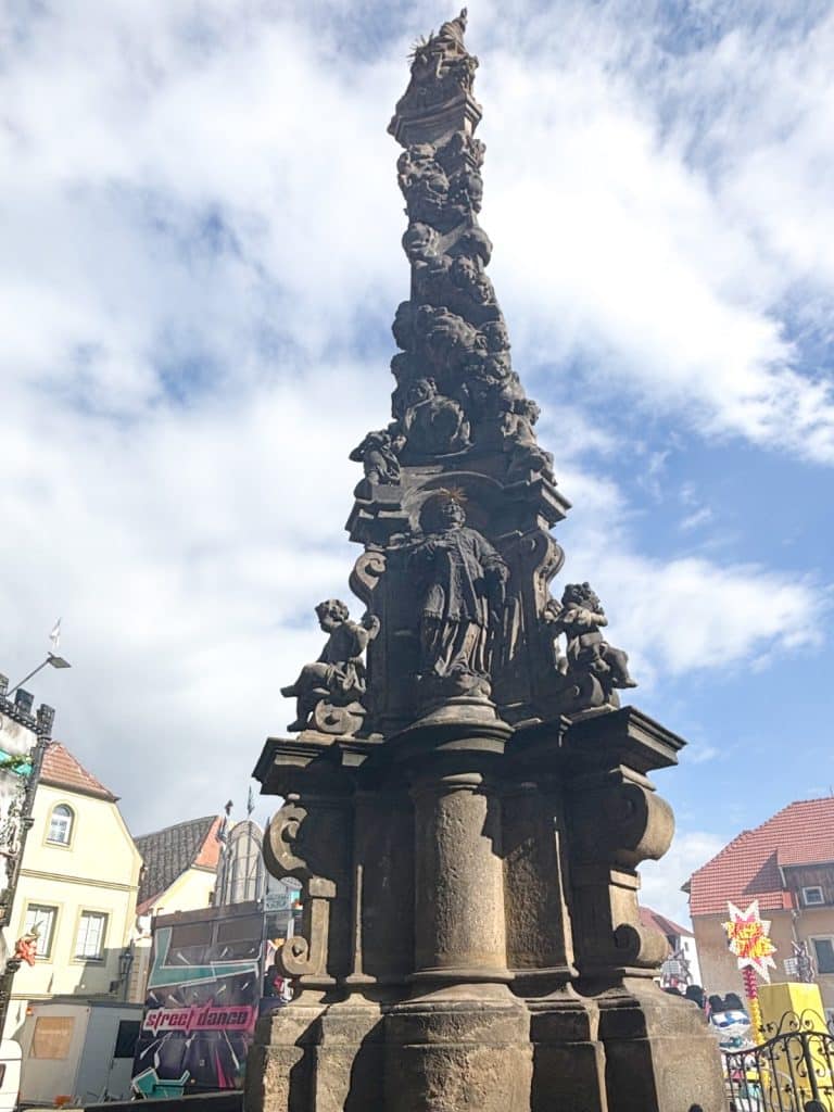 Eine detailreiche, barocke Pestsäule in Žatec aus dunklem Stein ragt vor einem leicht bewölkten blauen Himmel empor und zeigt zahlreiche Skulpturen von Heiligen und Engeln. Im Hintergrund sind Teile des Marktplatzes mit historischen Häusern und modernen Veranstaltungsbauten zu sehen.