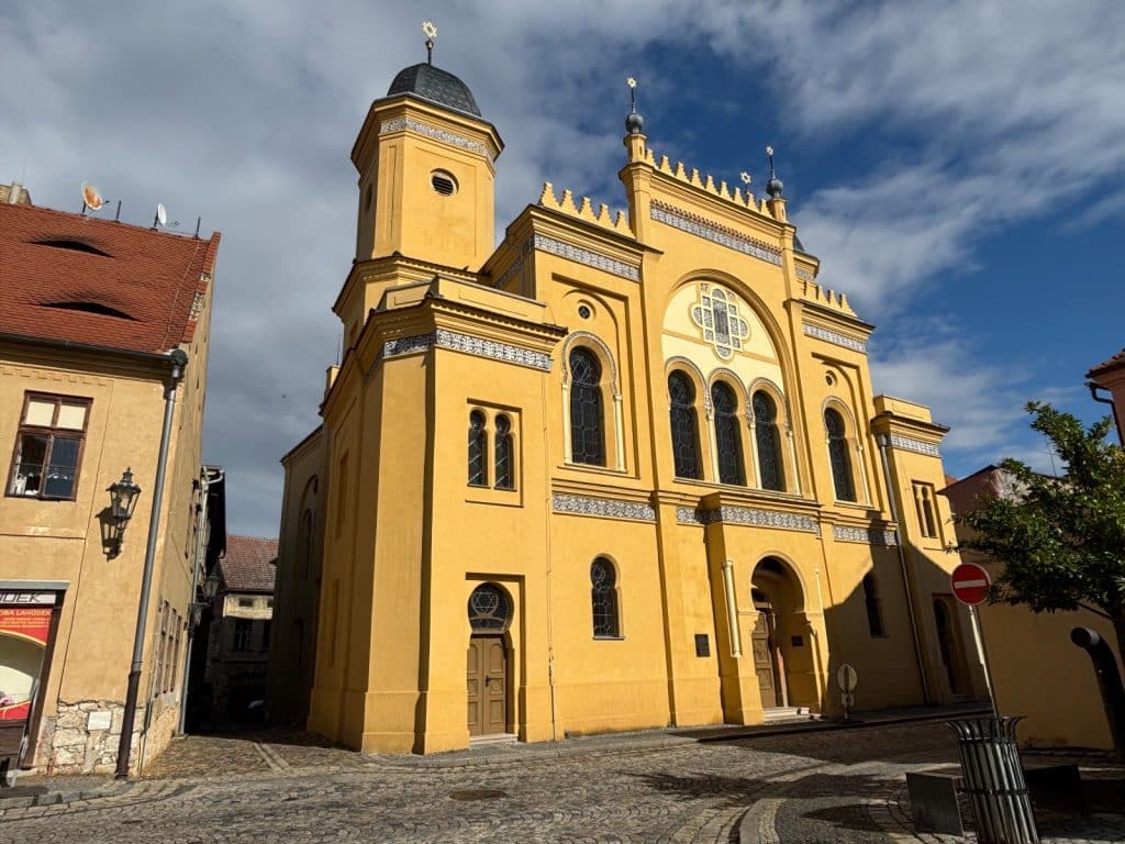 Die große Synagoge in Žatec präsentiert sich als imposantes, gelbes Gebäude im maurischen Stil mit markanten Türmen, Hufeisenbögen und Davidsternen unter einem blau-weißen Wolkenhimmel.