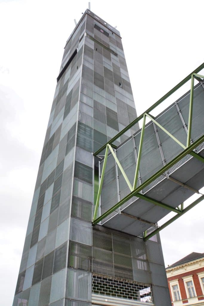 Blick von unten auf den modernen Zatrec Hopfenturm mit seiner markanten Fassade aus grauen Gitterelementen und einem angrenzenden, grünen Verbindungsgang aus Stahl.
