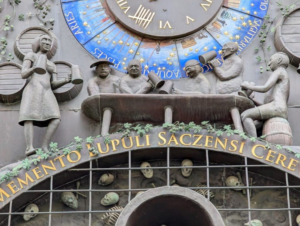 Die Zatrec Hopfenuhr zeigt eine Figurengruppe aus Bronze, die an einem Tisch Bier trinkt, unterhalb eines astrologischen Ziffernblatts und oberhalb eines Gitters mit Totenköpfen. Über den Figuren prangt ein goldener lateinischer Schriftzug, der von Hopfenranken umrahmt wird.