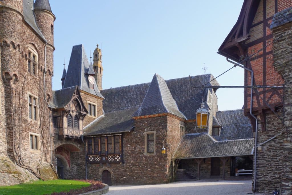 Blick in den gepflasterten Innenhof der Reichsburg Cochem mit historischen Gebäuden aus Bruchsteinmauerwerk, Fachwerkakzenten und verschiedenen Türmen unter einem klaren blauen Himmel.