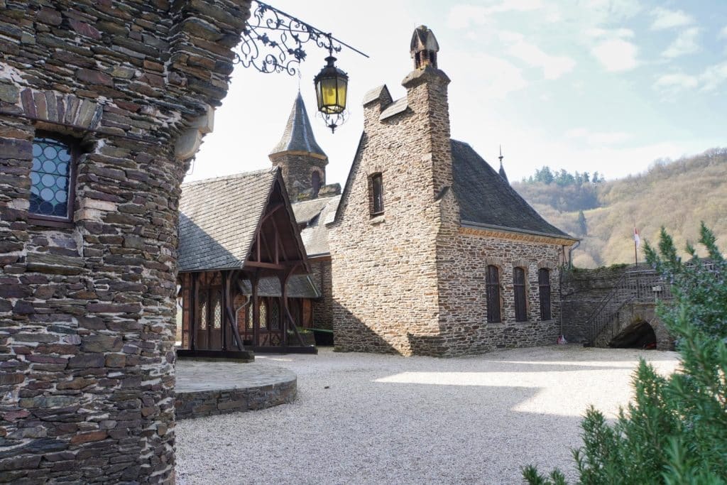 Der gepflasterte Innenhof der Reichsburg Cochem zeigt historische, aus Bruchsteinen errichtete Gebäude mit spitzen Dächern, Türmen und einer schmiedeeisernen Laterne im Vordergrund. Im Hintergrund erstreckt sich ein bewaldeter Hang unter einem hellen Himmel.