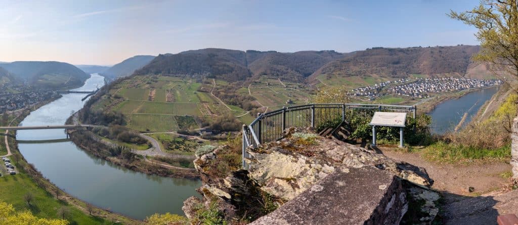 Panorama-Blick vom Aussichtspunkt in Neef auf eine weite Moselschleife mit Weinbergen, einer Brücke und dem gegenüberliegenden Ort unter einem klaren Himmel.