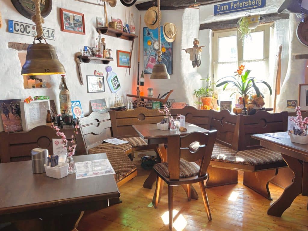 Der gemütliche Innenraum der Neef Einkehrstation ist mit rustikalen Holzmöbeln, karierten Sitzpolstern und zahlreichen nostalgischen Dekorationsgegenständen an den weißen Wänden ausgestattet. Ein sonnendurchflutetes Fenster beleuchtet die hölzerne Eckbank und die eingedeckten Tische, die mit kleinen Blumengestecken dekoriert sind.