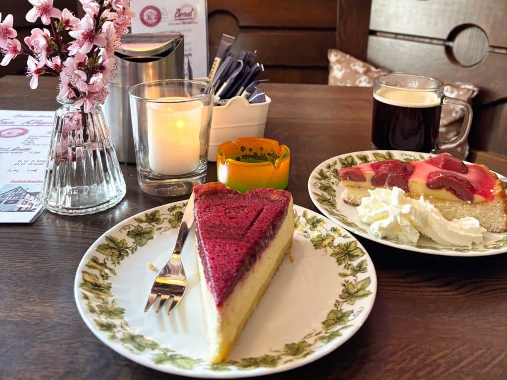 Zwei Stücke Kuchen auf gemusterten Tellern, eine Tasse Kaffee und eine Vase mit rosa Blüten stehen auf einem Holztisch in der Neef Einkehrstation.