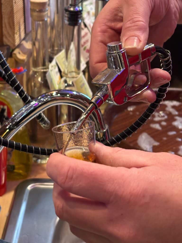 Nahaufnahme einer Person bei einem Tasting in der Neef Einkehrstation, die mit einer Miniatur-Zapfpistole eine bernsteinfarbene Flüssigkeit in ein kleines Glas füllt.