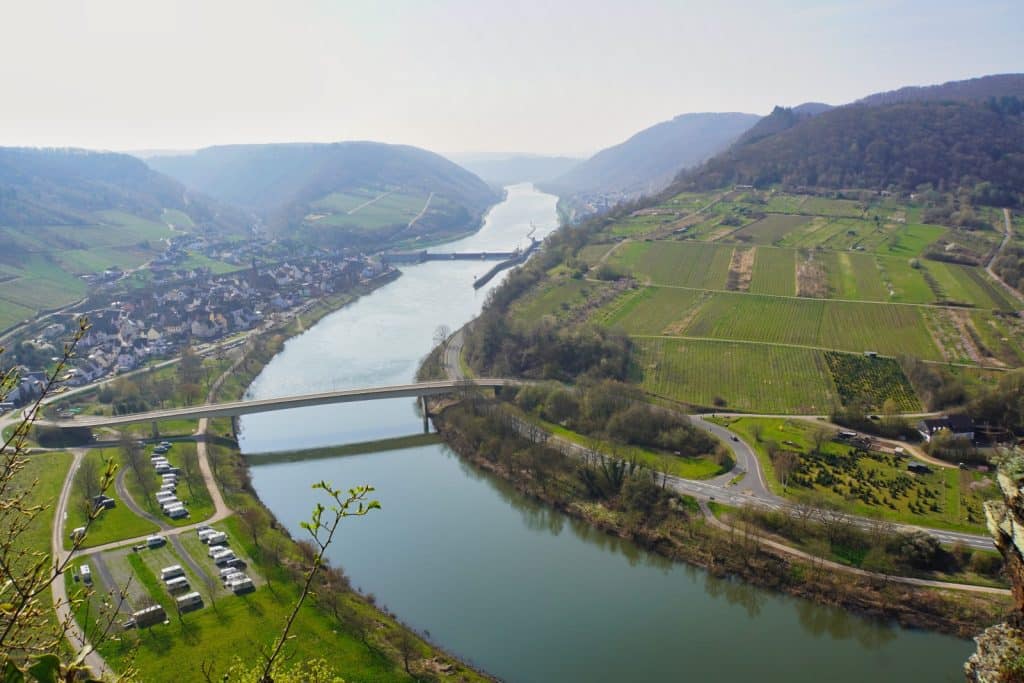 Ein weiter Blick von oben auf die Moselschleife bei Neef mit einer Brücke über den Fluss, einem Wohnmobilstellplatz am Ufer und Weinbergen an den umliegenden Hängen.