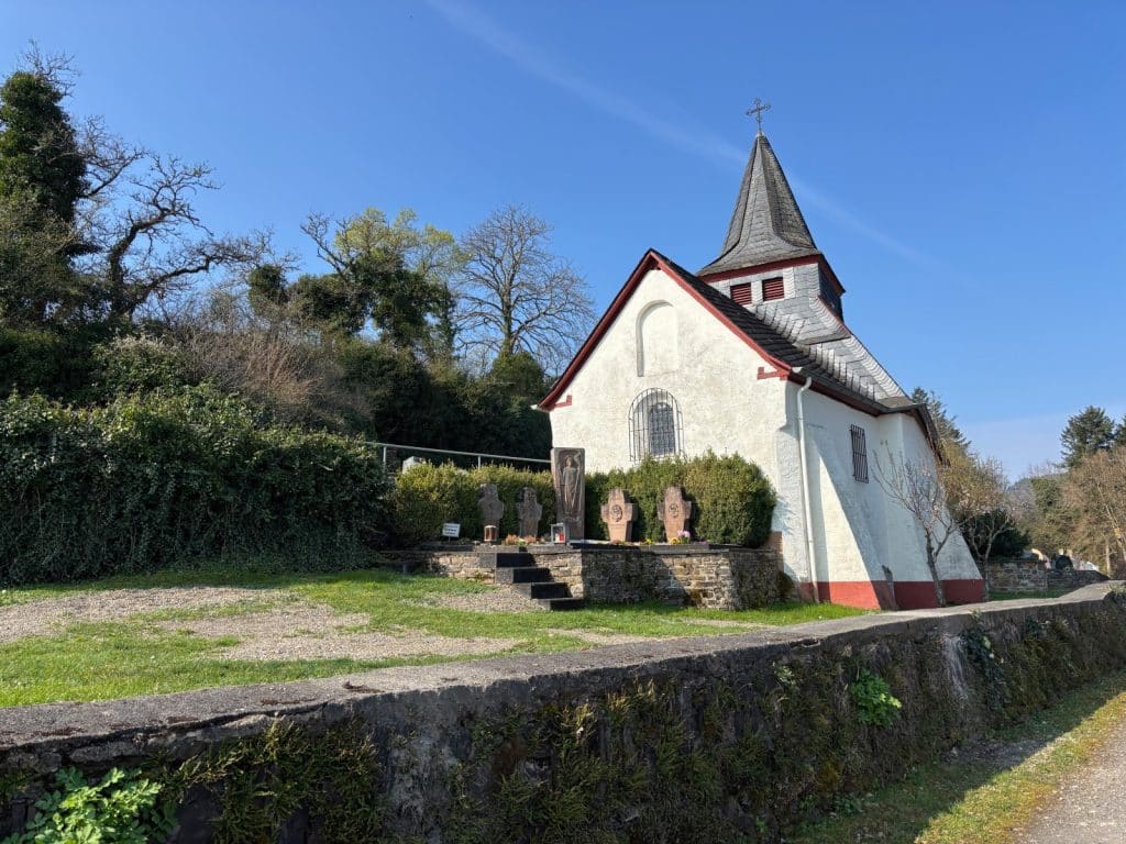 Die kleine, weiß verputzte Peterskapelle in Neef steht an einem Hang mit angrenzenden Grabkreuzen unter strahlend blauem Himmel.