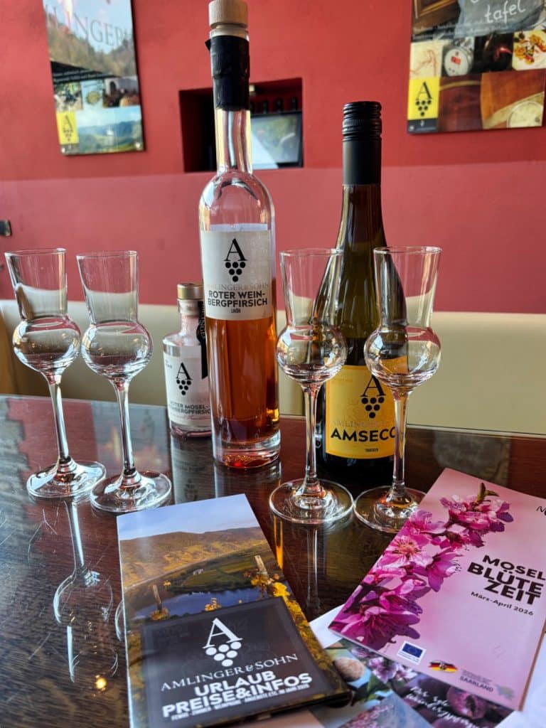 Auf einem Holztisch im Weingut Amlinger & Sohn in Neef stehen zwei Weinflaschen, ein kleiner Flachmann und vier Grappagläser neben Informationsbroschüren zur Mosel-Region. Die Flaschen tragen Etiketten mit der Aufschrift „Roter Weinbergpfirsich“ und „Amsecco“, während im Vordergrund ein Prospekt für Urlaub an der Mosel liegt.