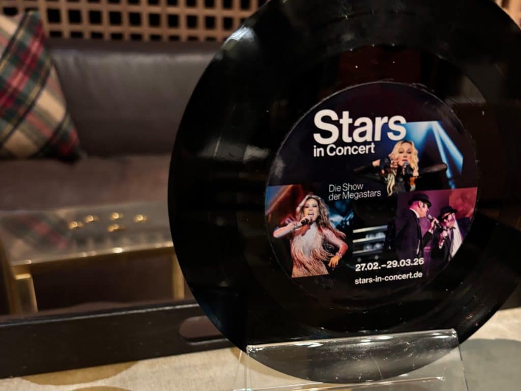 Eine Nahaufnahme zeigt eine kleine Schallplatte in einem Aufsteller, die als Werbeartikel für die Show „Stars in Concert“ in Berlin dient und Fotos von verschiedenen Doppelgängern sowie Veranstaltungsdaten zeigt. Im Hintergrund ist ein unscharfes Interieur mit einem Sofa, einem karierten Kissen und einem Couchtisch zu sehen.