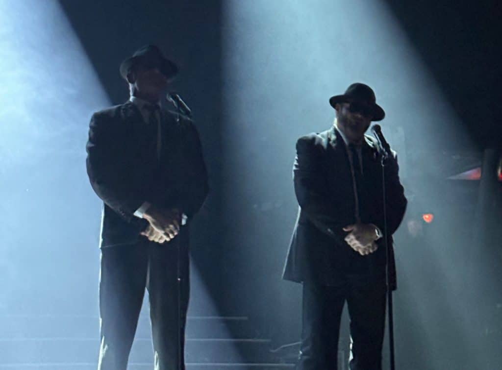 Zwei Darsteller der Show „Stars in Concert“ stehen als Blues Brothers in schwarzen Anzügen und Hüten unter hellen Scheinwerferkegeln auf einer dunklen Bühne in Berlin.