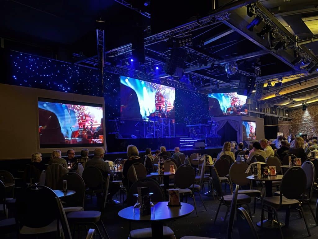 Blick von den Zuschauerplätzen auf die hell erleuchtete Bühne der Show „Stars in Concert“ in Berlin, die mit großen LED-Leinwänden und einer sternenähnlichen Hintergrundbeleuchtung ausgestattet ist.