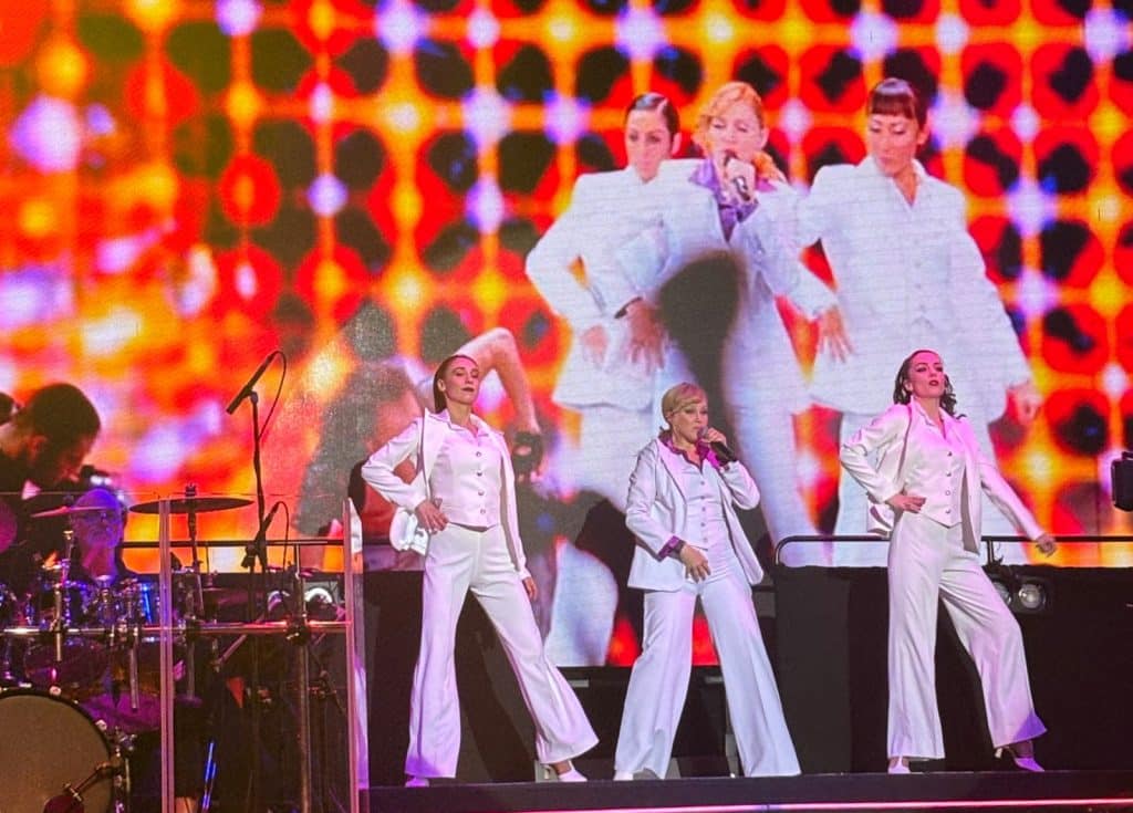 Drei Frauen in weißen Anzügen treten bei der Show „Stars in Concert“ in Berlin als Madonna-Double und Backgroundtänzerinnen vor einer großen LED-Leinwand auf, die ein Video der echten Madonna zeigt.