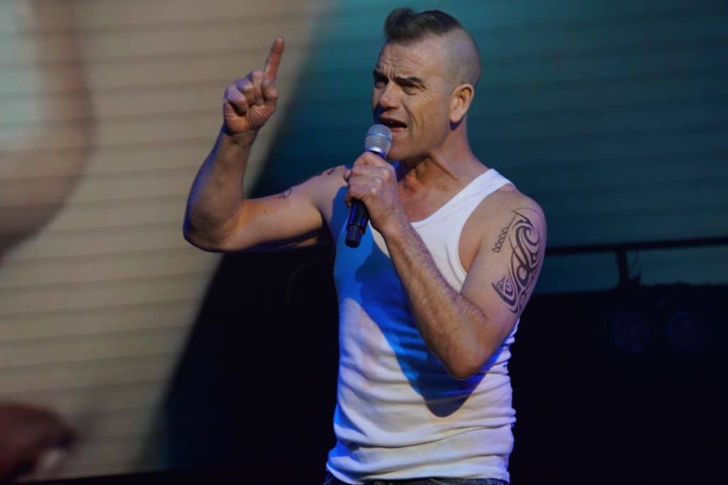 Ein Sänger tritt als Robbie-Williams-Imitator bei der Show „Stars in Concert“ in Berlin auf, trägt ein weißes Unterhemd und hält ein Mikrofon, während er mit erhobenem Zeigefinger singt.