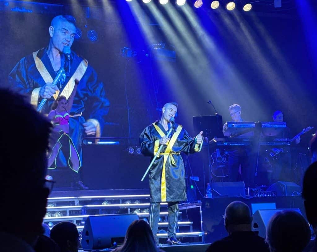 Ein Robbie-Williams-Imitator performt bei „Stars in Concert“ in Berlin in einem schwarz-goldenen Boxermantel auf einer blau beleuchteten Bühne, während im Hintergrund eine Großaufnahme von ihm projiziert wird.
