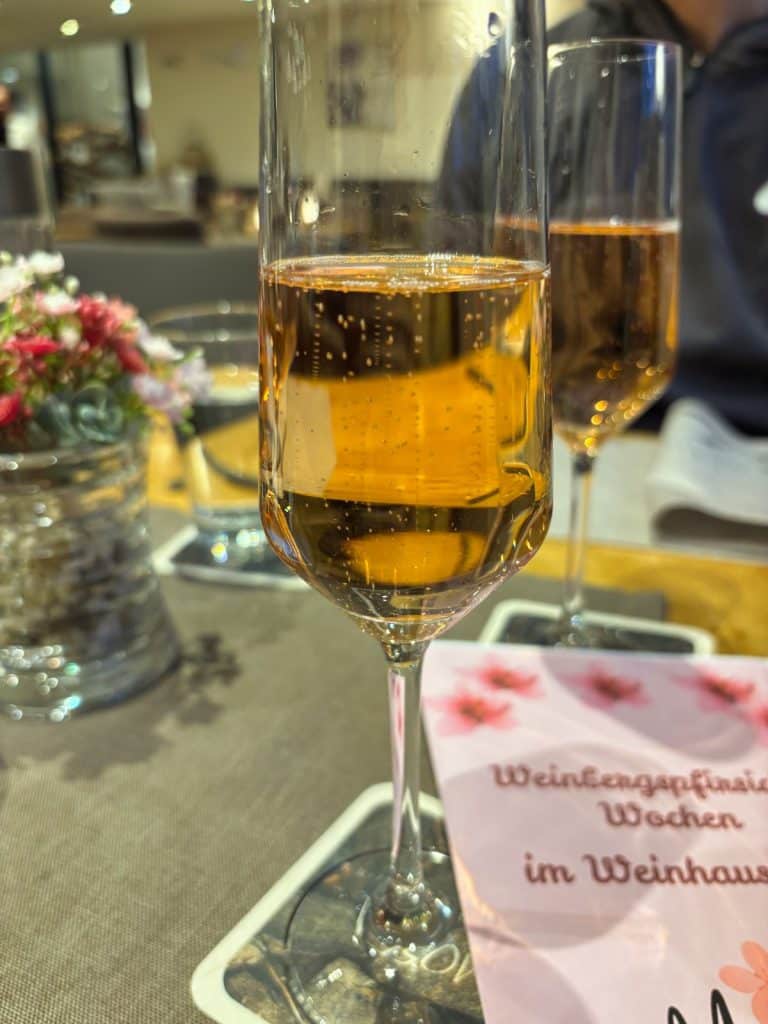 Ein goldfarbener Aperitif mit feinen Kohlensäurebläschen in einem Sektglas steht auf einem Tisch im Weinhaus in Bremm, im Hintergrund ist ein zweites Glas und Blumenschmuck zu sehen.