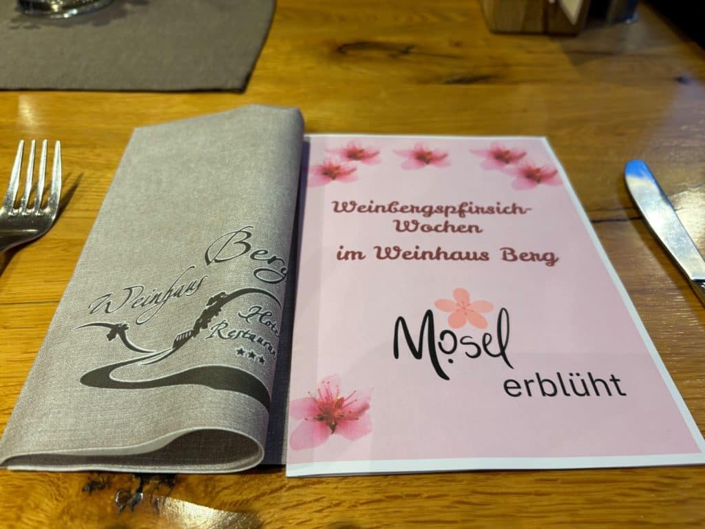Eine Speisekarte im Restaurant Weinhaus Berg in Bremm mit dem Titel „Weinbergpfirsich-Wochen“ und dem Schriftzug „Mosel erblüht“, dekoriert mit rosa Blüten neben einer grauen Stoffserviette.