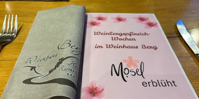 Eine Speisekarte im Restaurant Weinhaus Berg in Bremm mit dem Titel „Weinbergpfirsich-Wochen“ und dem Schriftzug „Mosel erblüht“, dekoriert mit rosa Blüten neben einer grauen Stoffserviette.