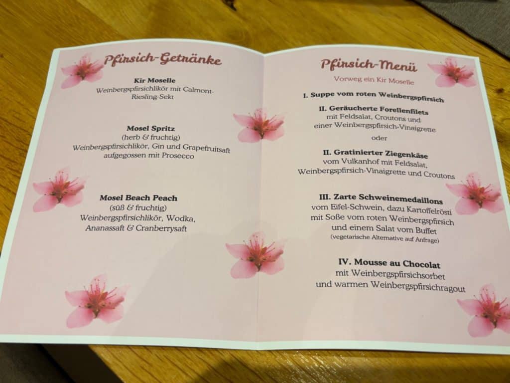 Ein zweigeteiltes Menü aus Bremm zeigt links „Pfirsich-Getränke“ wie den Kir Moselle und rechts eine viergängige „Pfirsich-Menüabfolge“, die unter anderem Suppe vom roten Weinbergpfirsich, Schweinemedaillons und Mousse au Chocolat umfasst. Das weiße Papier ist mit zartrosa Pfirsichblüten illustriert und liegt auf einem hellen Holztisch.
