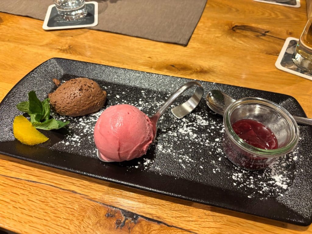 Eine Nachspeise im Restaurant in Bremm, bestehend aus einer Kugel Schokoladenmousse, einer Kugel rosa Sorbet auf einem Servierlöffel und einem kleinen Glas Beerenkompott, angerichtet auf einer schwarzen Platte mit Puderzucker, Minze und einer Orangenspalte.