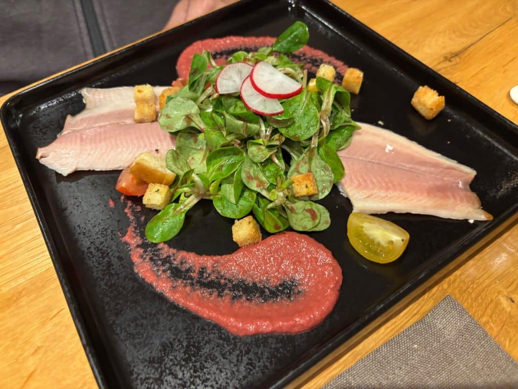 Ein modern angerichteter Forellensalat in einem Restaurant in Bremm mit zwei hellen Fischfilets, einem zentralen Haufen Feldsalat mit Radieschenscheiben, Croutons und zwei geschwungenen Streifen roter Sauce auf einem quadratischen schwarzen Teller.