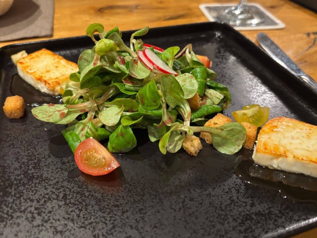 Ein modern angerichteter Salat mit Feldsalat, Radieschenscheiben, Kirschtomaten und Croutons, serviert mit zwei Stücken gebratenem Ziegenkäse auf einem flachen, schwarzen Teller im Bremm Restaurant.