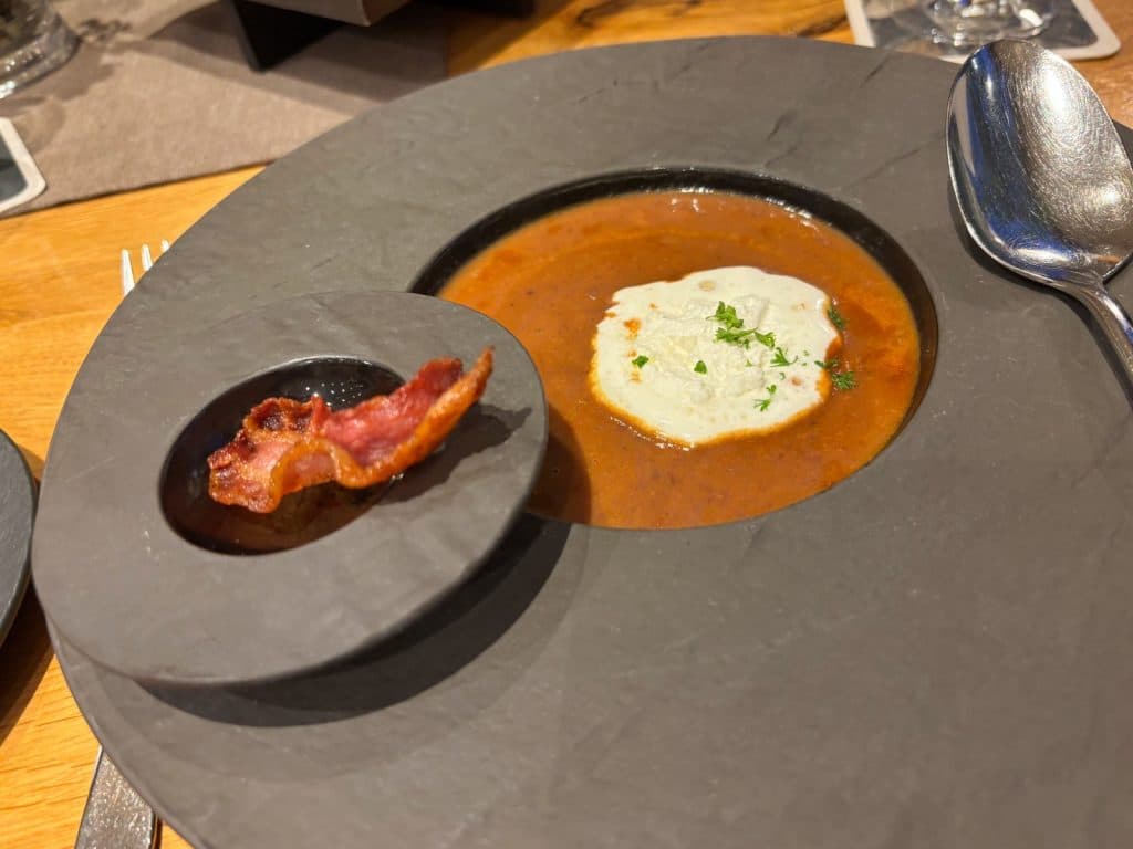 Eine cremige, rötliche Suppe in einem modernen, schiefergrauen Teller, garniert mit einem Klecks Sahne und Kräutern sowie einem knusprigen Speckstreifen auf einer separaten Vertiefung im Tellerrand.