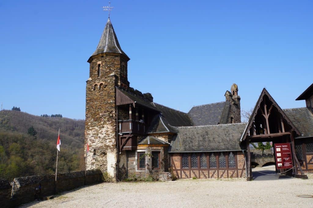 Der historische Burghof der Reichsburg Cochem zeigt einen markanten Steinturm mit spitzem Dach, angrenzende Fachwerkgebäude und ein hölzernes Eingangstor unter strahlend blauem Himmel. Im Hintergrund erstrecken sich bewaldete Hänge, während im Vordergrund ein heller Kiesplatz und eine niedrige Steinmauer zu sehen sind.